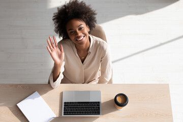 Smiling young woman waving hello while sitting at desk with laptop and coffee cup in bright modern office