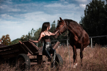 Obraz premium Sexy beautiful young woman in leather clothes petting a horse while sitting on wooden vintage cart in the field. Freedom and imprisonment. Friendship and loyalty. Peace of mind. Dramatic and nostalgic