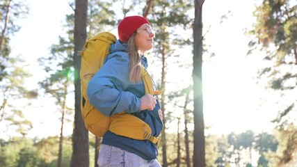 Woman hiking through sunlit pine forest wearing beanie backpack exploring nature trail sunlight filtering through tree creating warm glow outdoor adventure hiker vibe with calm travel spirit