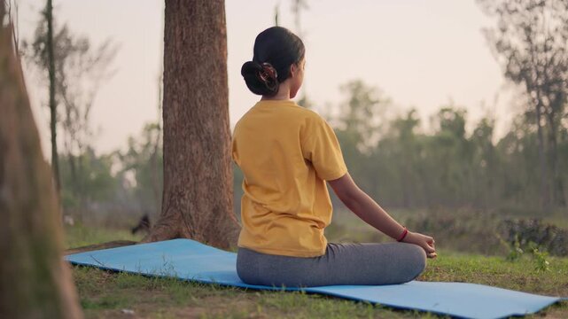 Back view of an indian woman sitting in padmasana pose on a yoga mat in the forest during golden hour.