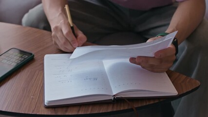 Handheld shot of young male Asian entrepreneur doing calculations with financial papers and taking notes in home office