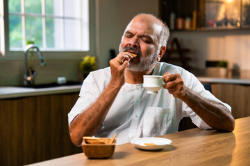 Elderly Indian man dips biscuit in tea at home kitchen