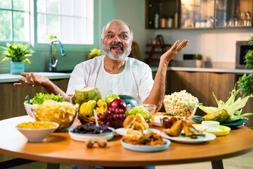 Happy senior Indian foodie enjoying snack-filled table in stylish kitchen setup