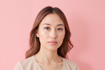 Portrait of young adult Asian woman looking directly at camera with neutral expression, long brown hair framing face, standing against plain pink background, head and shoulders visible