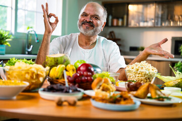 Happy senior Indian foodie enjoying snack-filled table in stylish kitchen setup