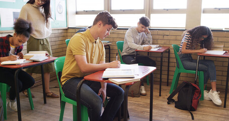 Exam, high school and teacher walking in classroom for assessment, test and quiz supervision. Teaching, academy and woman with students writing assignment for education, learning and lesson progress