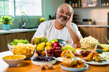 Happy senior Indian foodie enjoying snack-filled table in stylish kitchen setup