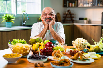 Happy senior Indian foodie enjoying snack-filled table in stylish kitchen setup