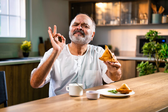 Happy senior Indian man enjoying samosa and tea at home in modern kitchen setting - Powered by Adobe
