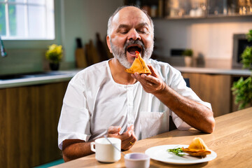Happy senior Indian man enjoying samosa and tea at home in modern kitchen setting