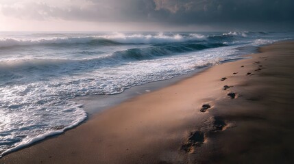 Obraz premium Footprints on a sandy beach with ocean waves gently breaking on the shore under a dramatic cloudy sky at sunrise evoking a peaceful journey