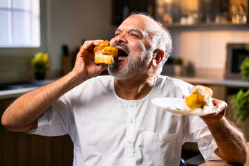 Elderly Indian senior foodie relishing Mumbai vada pav in modern Indian kitchen