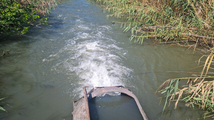 Rusty metal pipe discharging water into river with reeds and vegetation on banks. Industrial wastewater and environmental pollution concept