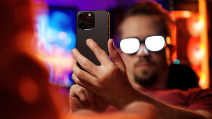 Man with anime glowing glasses swiping on smartphone relaxing in cyberspace home office laying on...