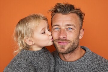 Obraz premium A young girl lovingly kisses her father's cheek against a warm, vibrant orange backdrop.