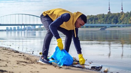 African American man volunteer gathers trash in blue bag, guy wearing yellow gloves picks up rubbish cleaning city beach near water, environmental protection concept, eco sustainability community clea - Powered by Adobe