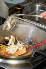 Chef adds Korean rice cakes to wok with vegetables and sauce in professional kitchen. Cooking tteokbokki or stir-fry. Close-up of hands, utensils, and vibrant ingredients