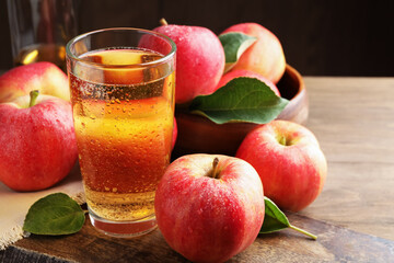 Delicious cider in glass, apples and leaves on wooden table against dark background, closeup