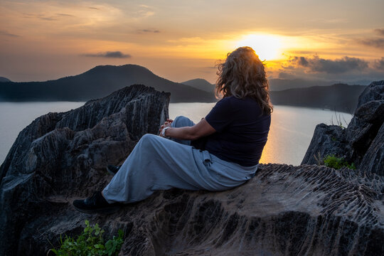 Woman sitting on rocks at sunset overlooking Valle de Bravo lake in Mexico from La Pena, symbolizing reflection, serenity, meditation, tourism and cultural identity