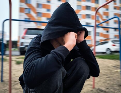 sad lonely teenager. guy in a black hoodie. teenage boy in depression. courtyard of a multi-storey residential building, parked cars, and a teenage boy alone on street