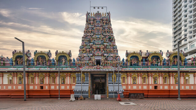 Kandaswamy Kovil Hindu temple, Kuala Lumpur, Malaysia. Intricate sculptures adorn the traditional architecture, contrasting with a modern high-rise under an overcast sky