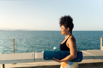 Curvy woman walking on pier by sea carrying a yoga mat and water bottle, a healthy lifestyle