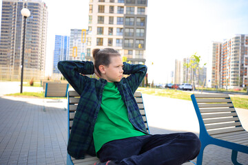 a teenager sitting alone on a bench, looking bored amidst the imposing of the city on a summer day. urban environment and the teenager's expression, isolation and longing for connection.