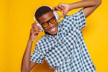 Young afro latin american man enjoying music with headphones and sunglasses, broadly smiles on yellow background.
