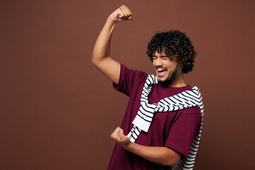 Young excited happy Indian man he wear purple t-shirt sweatshirt tied over shoulders casual clothes doing winner gesture celebrate clenching fists isolated on plain brown background Lifestyle concept