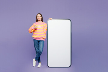 Full body young smiling woman wears yellow sweatshirt casual clothes big huge blank screen mobile cell phone smartphone with area show thumb up isolated on plain purple background. Lifestyle concept.