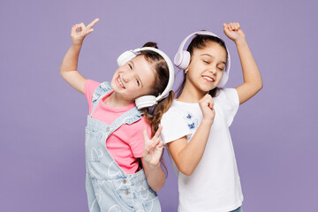 Little cheerful two friends kids children girls 9-10 years old wear casual clothes together listen to music in headphones raise up hands dance isolated on plain purple background. Lifestyle concept.