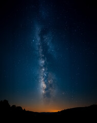Fototapeta premium Stunning vertical shot of the Milky Way illuminating the night sky above a dark mountainous horizon, with a warm afterglow rising from the bottom of the frame.