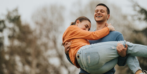 A cheerful young couple enjoys a playful moment together in nature. The man lifts the smiling woman in his arms, both displaying happiness and a sense of togetherness on a serene outdoor day.