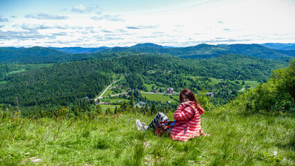 Fototapeta premium woman 30-40 years old sits in the grass, on a meadow, turns into the lens, high in the mountains, red paita, long brown hair, view of the mountains, houses, forest, clouds