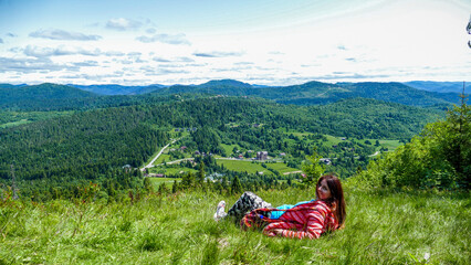 Fototapeta premium woman 30-40 years old lying in the grass, on a meadow, turning into the lens, high in the mountains, red coat, long brown hair, view of the mountains, houses, forest, clouds