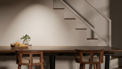 Breads and glass of coffee on wooden dining table and chairs beside stairs in dim lit room or house.