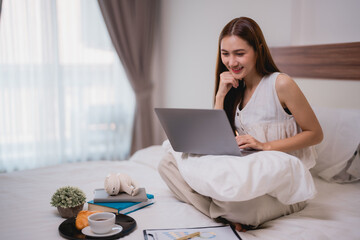 Asian businesswoman working from home, relaxing in bed with breakfast while using a laptop. Smiling and engaged with the computer screen, enjoying a productive morning