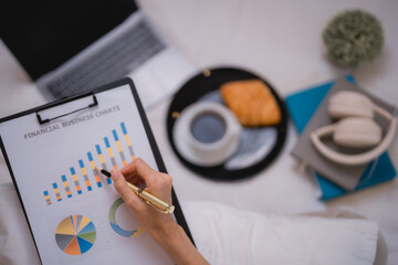 Businesswoman pointing at financial business charts with a pen, drinking coffee and eating a croissant, working from home or during a relaxing break at the office