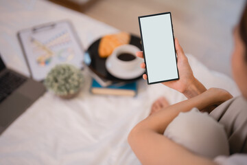 Relaxing on a bed, a woman holds a smartphone with a blank screen, surrounded by a laptop, coffee, and a croissant, creating an inviting scene perfect for mockups
