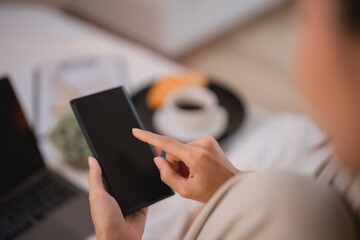 Businesswoman using smartphone with blank screen while having breakfast in bed, with a laptop, croissant and coffee on a tray in the background