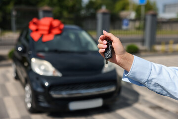 Dreams come true. Woman holding car key near her new auto with big red bow outdoors, closeup