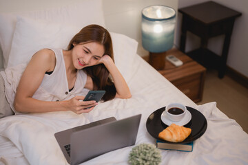 Smiling young woman lies comfortably in bed, using her smartphone and laptop while enjoying a breakfast of coffee and croissants, suggesting a relaxing and productive start to her day