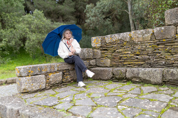Senior woman holding blue umbrella sitting on stone wall in park
