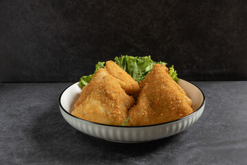 Risol Mayo segitiga or Triangle Risol mayonnaise served in a white bowl with fresh lettuce and green chili on dark background. Showcasing a savory Indonesian snack with breadcrumb coating. 