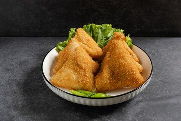 Risol Mayo segitiga or Triangle Risol mayonnaise served in a white bowl with fresh lettuce and green chili on dark background. Showcasing a savory Indonesian snack with breadcrumb coating. 