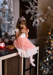 A girl in a pink dress sits by herself at a table, sipping cocoa beside a Christmas tree.