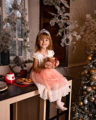 A little girl in a pink dress sits on a table next to a Christmas tree in a cozy New Year's atmosphere.