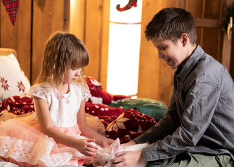 Brother and sister sit on a red plaid blanket, tenderly playing with a white rabbit. Christmas decorations are all around.