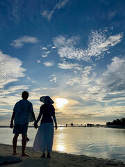 Fototapeta premium Silhouette of a couple standing side by side, holding hands and facing the ocean during a vibrant sunset, with colorful sky and beach scenery