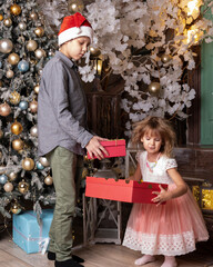 A family celebrates Christmas, a boy in a Santa hat and a girl in a fluffy dress hold gifts next to a Christmas tree.
